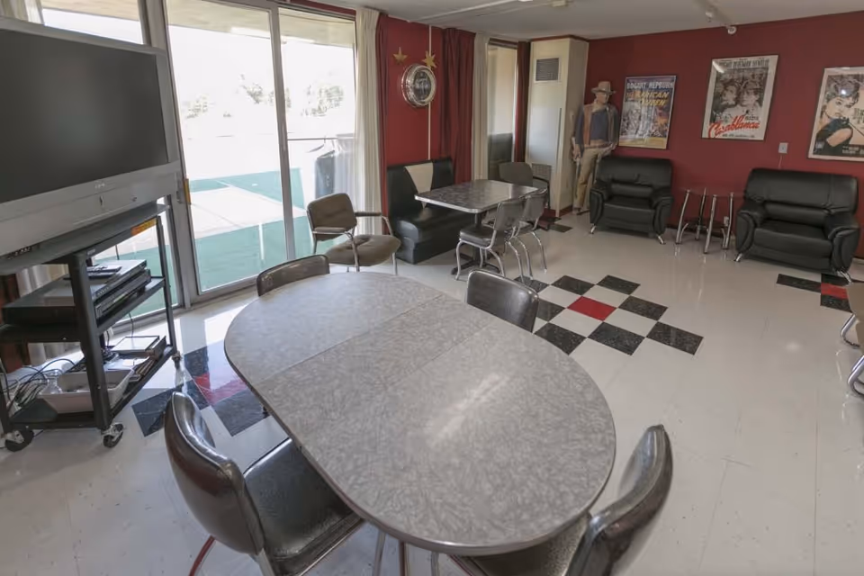 Community dining/activity room with oval and square tables, chairs, a TV on a cart, recliners, and sliding glass doors to the outside.