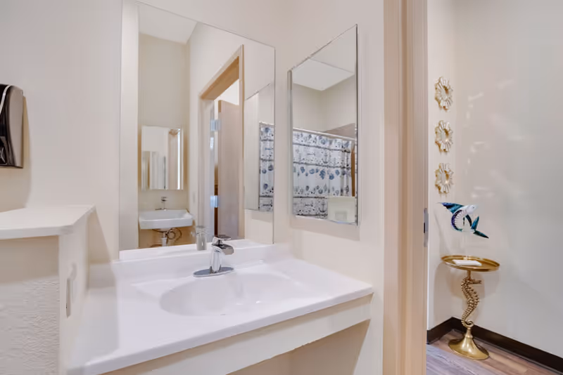 A clean and bright bathroom sink area with a white countertop and a modern faucet. Two mirrors are mounted on the walls above the sink, reflecting a small white sink and a shower curtain with a blue and white pattern in the adjacent room. To the right, there is a decorative gold stand with a blue and white fish sculpture and three gold flower wall decorations.
