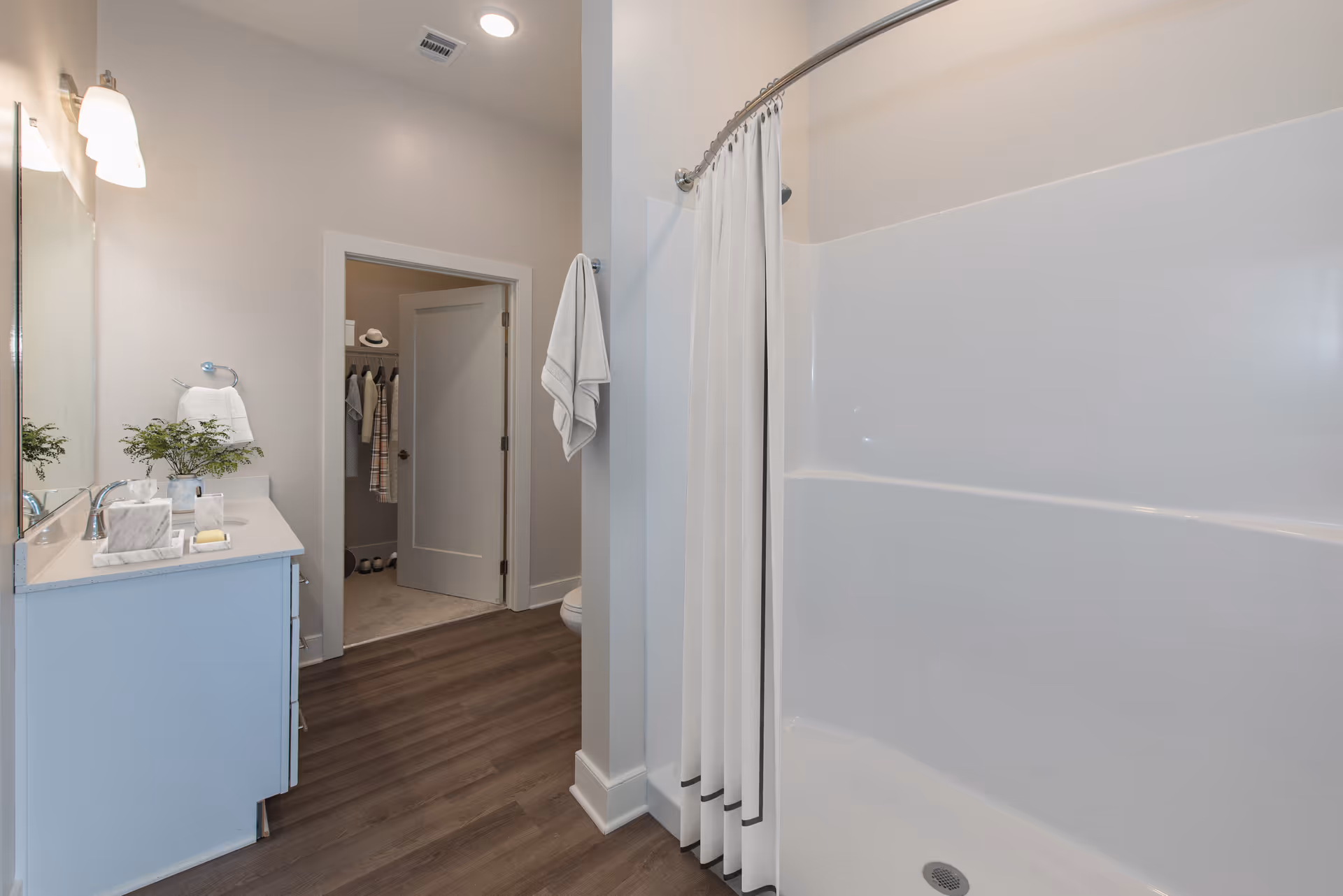 A clean and modern bathroom featuring a white vanity with a sink, a large mirror, and a light fixture above. There is a white shower with a curtain on the right side. A towel hangs on a hook on the wall near the shower. The bathroom has wood-look flooring and an open door leading to a closet with hanging clothes and shoes visible.