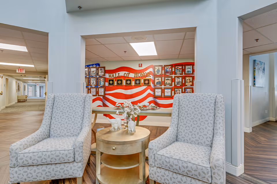 Two patterned armchairs with a round wooden table between them, holding a vase with flowers and decorative items, set in a senior living facility hallway. In the background, there is a Wall of Honor display with an American flag design and framed photos and plaques.