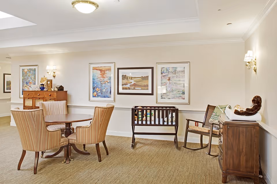 A cozy interior room with a round wooden table surrounded by four striped upholstered chairs. On the right side, there is a wooden baby crib with toys inside, a wooden rocking chair, and a wooden dresser with a changing pad and a stuffed monkey on top. The walls are decorated with three framed paintings and two wall sconces with lights.