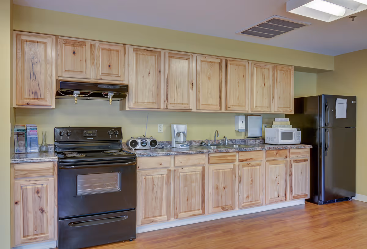 A kitchen area with wooden cabinets, a black stove and oven, a coffee maker, a small radio, a microwave, a sink, and a black refrigerator. The floor is wooden and the walls are painted light green.
