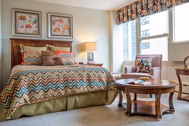 Sunlit furnished bedroom with a patterned bedspread, armchair, wooden coffee table, and large window.