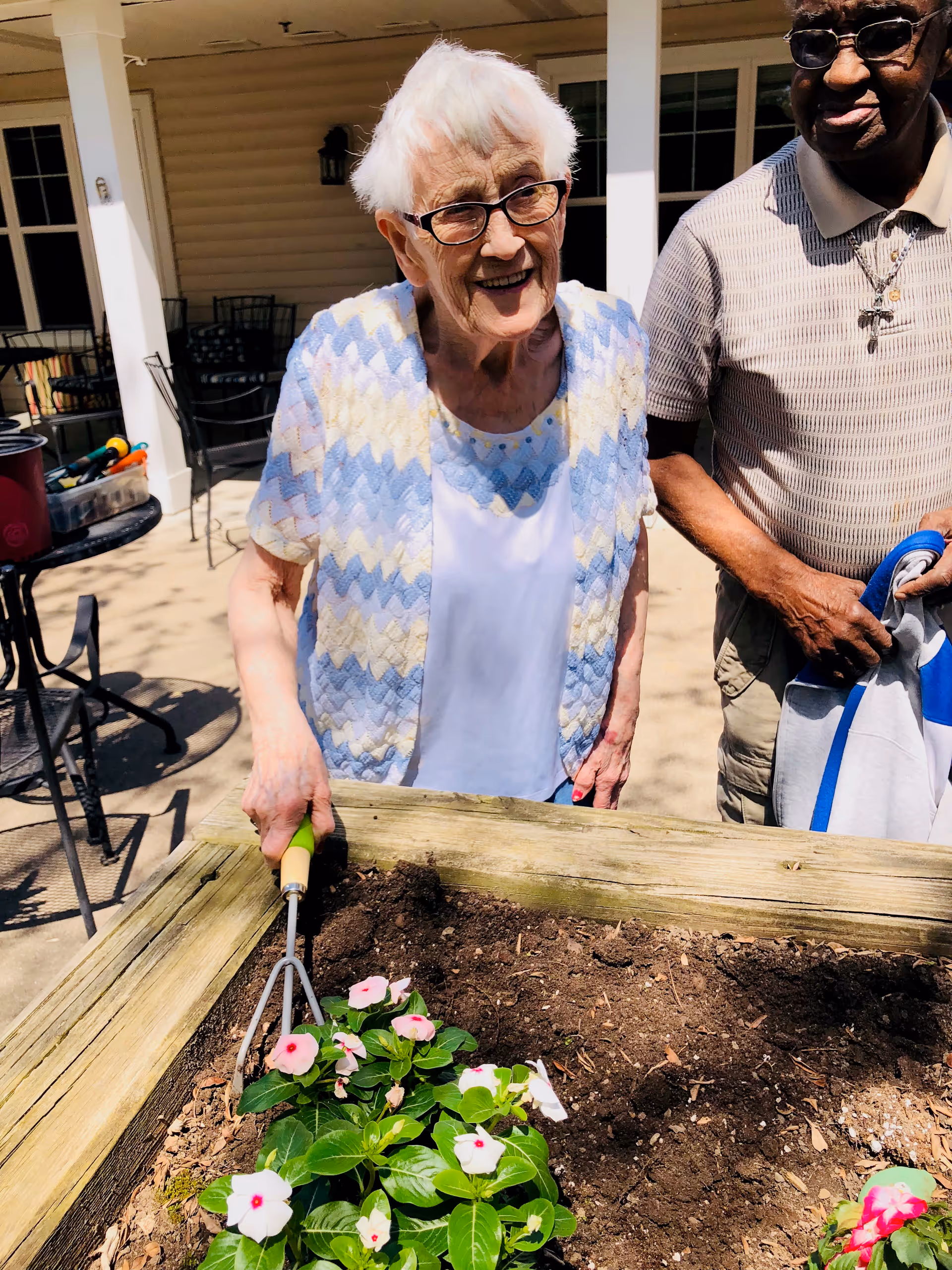 An elderly woman with white hair and glasses is smiling while gardening at a raised wooden flower bed with pink and white flowers. She is holding a small gardening tool. Next to her stands an elderly man wearing glasses, a striped polo shirt, and a necklace with a cross, holding a jacket. They are outside on a patio area with chairs and tables in the background.