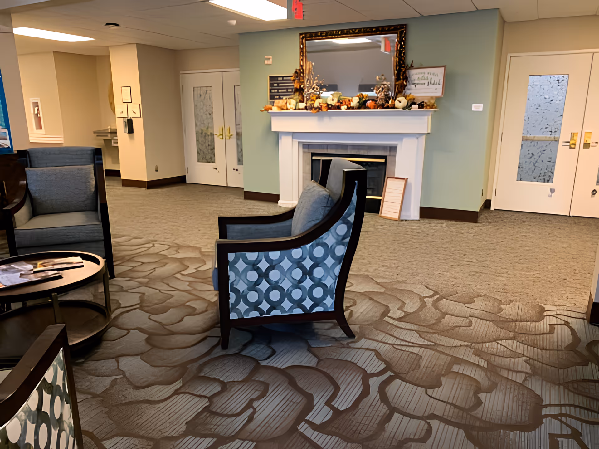 A cozy seating area in a senior living facility with patterned armchairs arranged around a small round table. The room features a decorative fireplace with a large mirror above it and seasonal decorations on the mantel. Double doors with frosted glass panels are visible in the background.