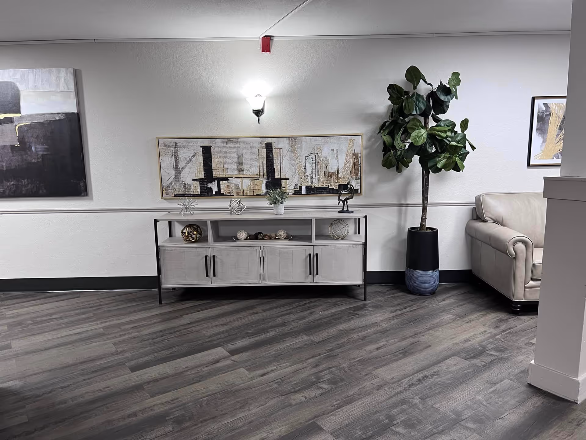A modern interior space with a light gray wooden floor, a beige couch on the right, a tall potted plant next to the couch, and a light gray console table against the wall. The console table has decorative items including geometric sculptures and a small plant. Above the console table is a rectangular abstract painting, and to the left on the wall is another abstract painting. A wall sconce light fixture is mounted above the console table.
