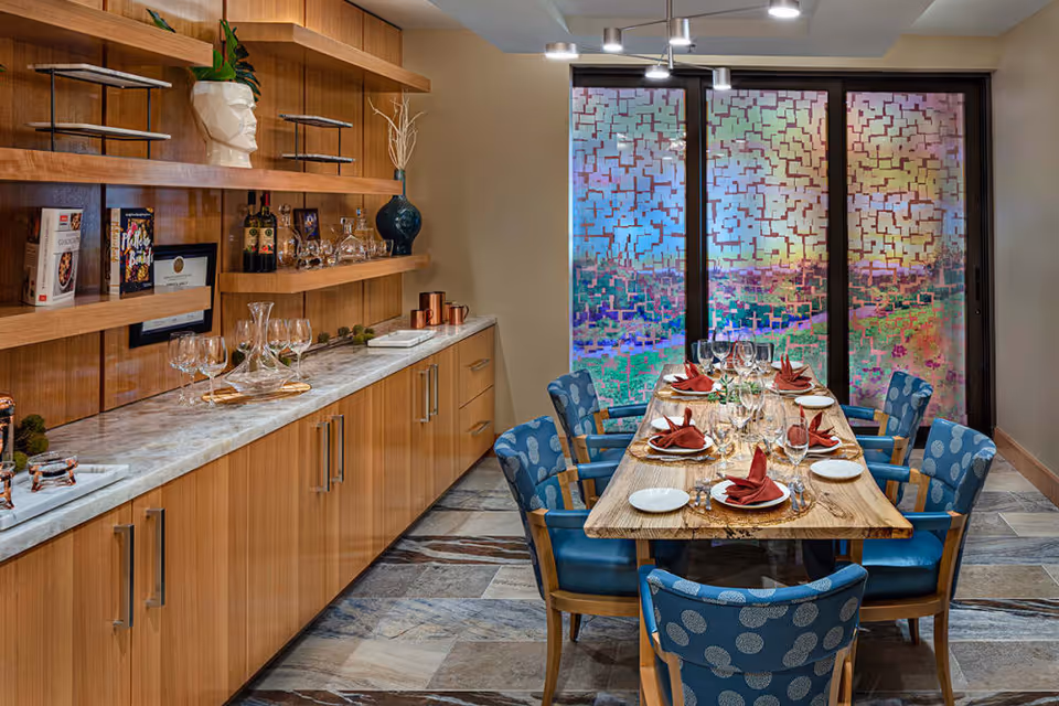 A dining room with a wooden table set for six people with plates, wine glasses, and red folded napkins. The chairs are blue with a circular pattern. Behind the table is a large window with a colorful, abstract patterned privacy film. On the left side, there is a wooden cabinet with marble countertop, decorated with glassware, bottles, books, and decorative items.