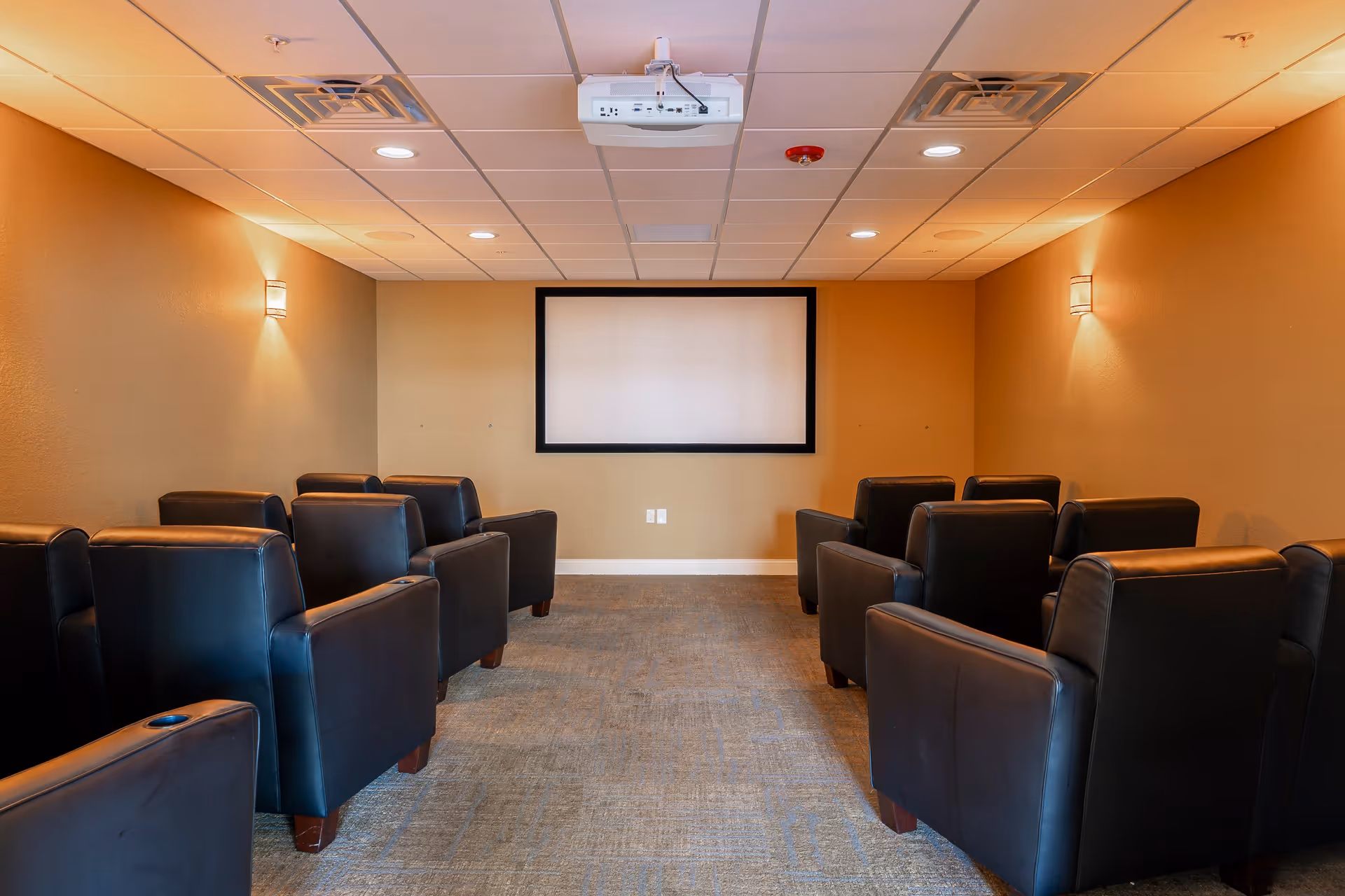 A small theater room with two rows of black leather armchairs facing a blank projection screen mounted on a beige wall. The ceiling has recessed lighting and a projector is mounted in the center. The walls are painted in warm tones with wall sconces providing soft lighting.
