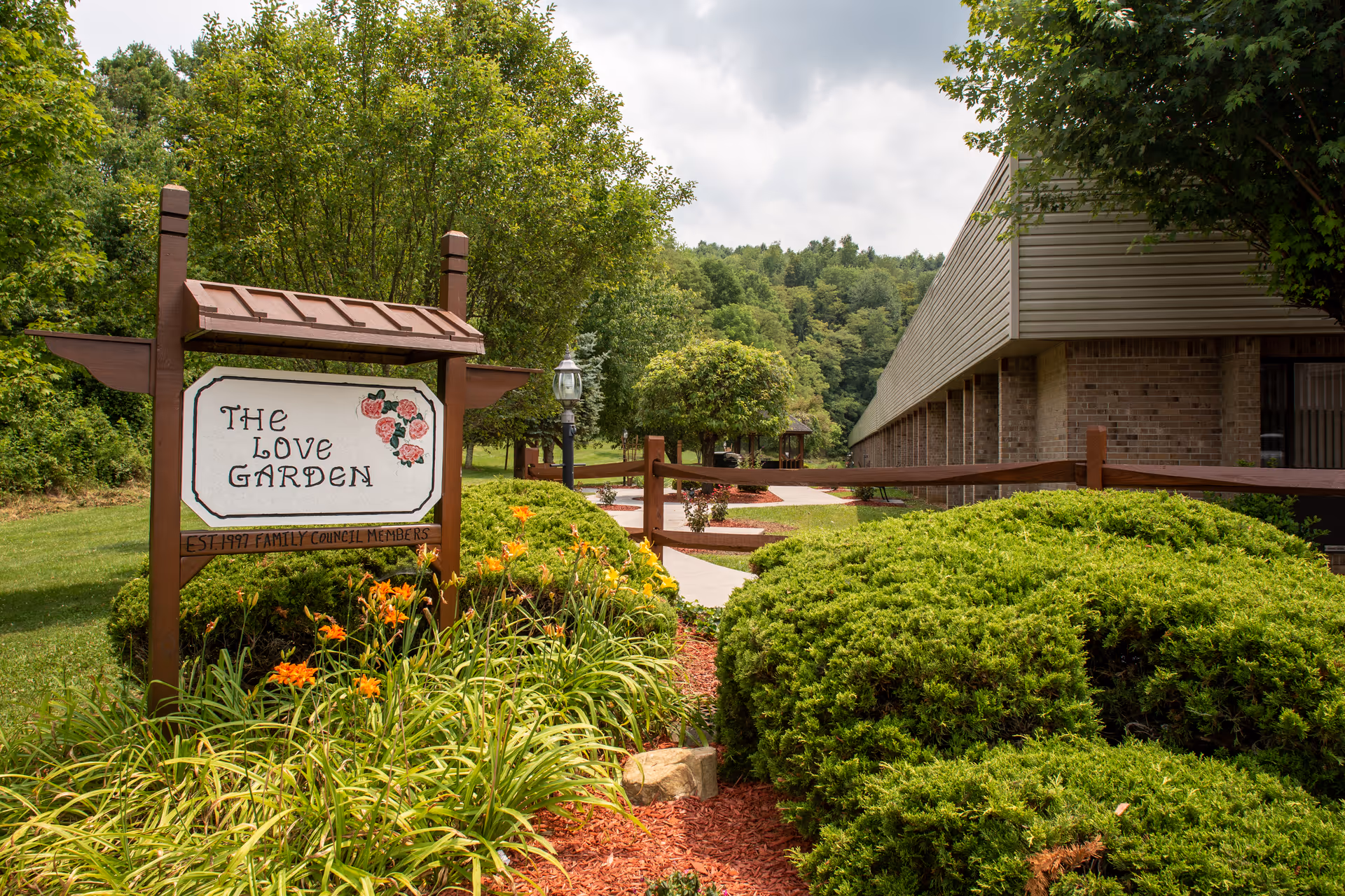 Landscaped outdoor garden with a wooden sign reading "The Love Garden", flowering plants, shrubs, a walkway, and a building in the background.
