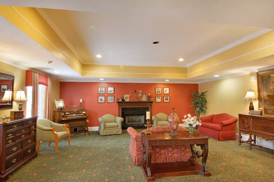 A warm communal living room with sofas and armchairs arranged around a central wooden coffee table and fireplace, decorated with lamps, framed pictures, and a patterned carpet.
