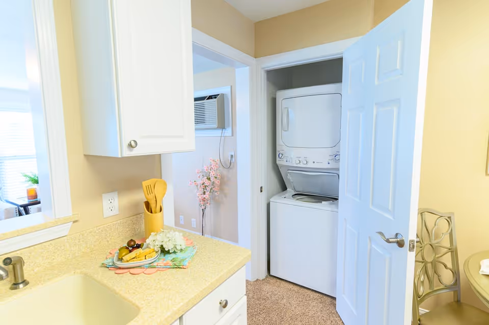 A bright kitchen area with a beige countertop and white cabinets. On the countertop, there is a plate with fruit and flowers, along with wooden kitchen utensils in a holder. To the right, there is an open door revealing a stacked washer and dryer unit in a small laundry closet. In the background, part of a living room with a window air conditioning unit and a vase with pink flowers is visible.