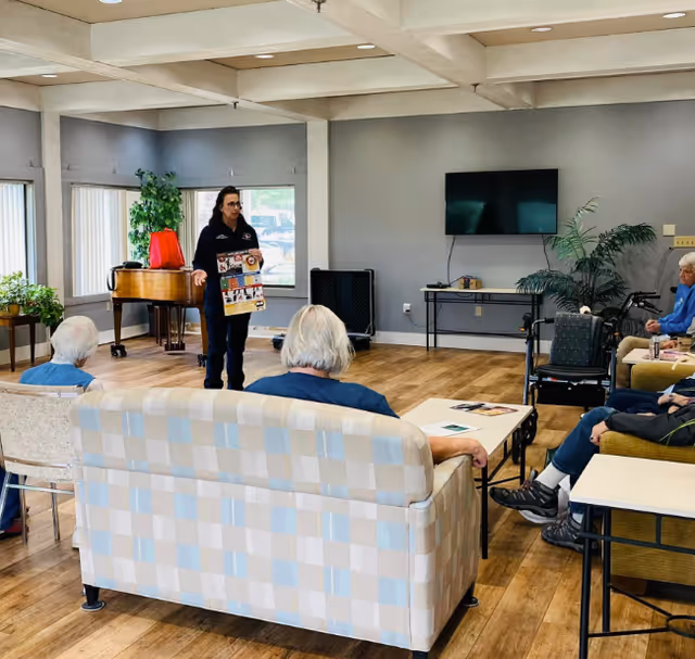 A group of elderly people seated in a common room with a woman standing and holding a poster, possibly giving a presentation. The room has wooden floors, a piano, a TV mounted on the wall, and some plants.