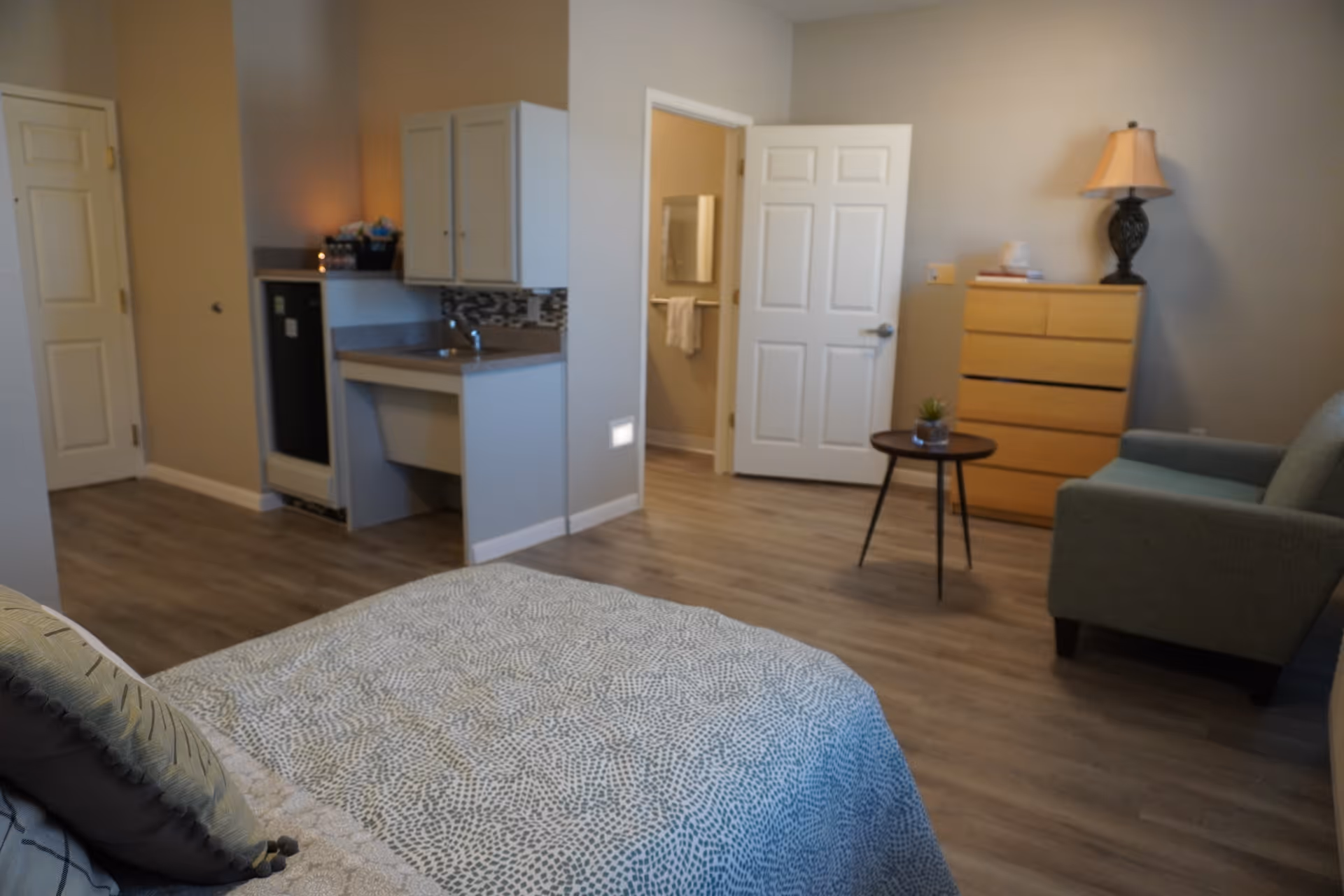 A cozy bedroom in a senior living facility featuring a bed with patterned bedding, a small kitchenette with a sink and mini fridge, a wooden dresser with a lamp and books on top, a small round table with a plant, and a comfortable armchair. The room has light-colored walls and wood flooring, with an open door leading to a bathroom.