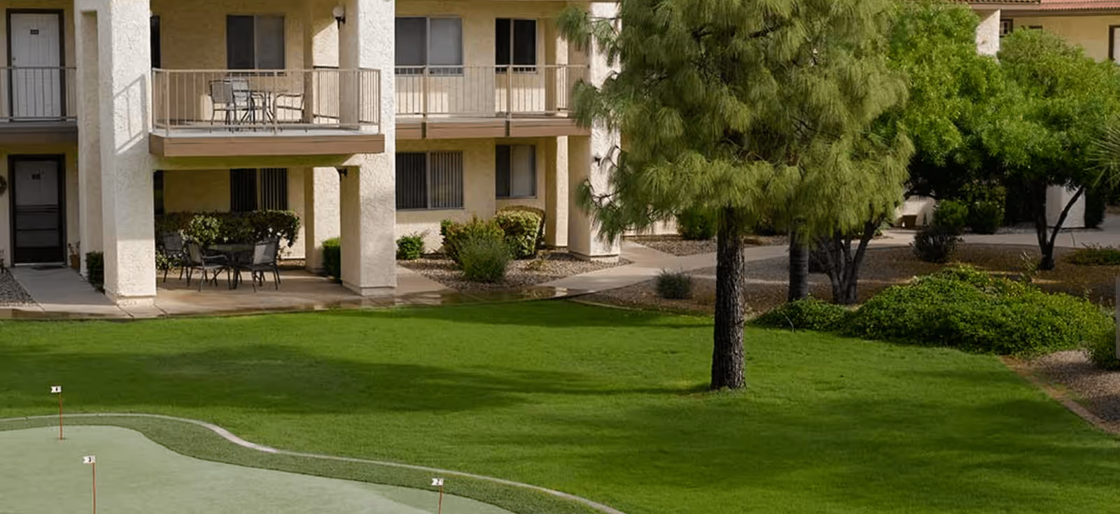 Outdoor view of a senior living facility showing a well-maintained green lawn with a putting green and small golf flags. The background features a two-story building with balconies and patio furniture, surrounded by trees and shrubs.