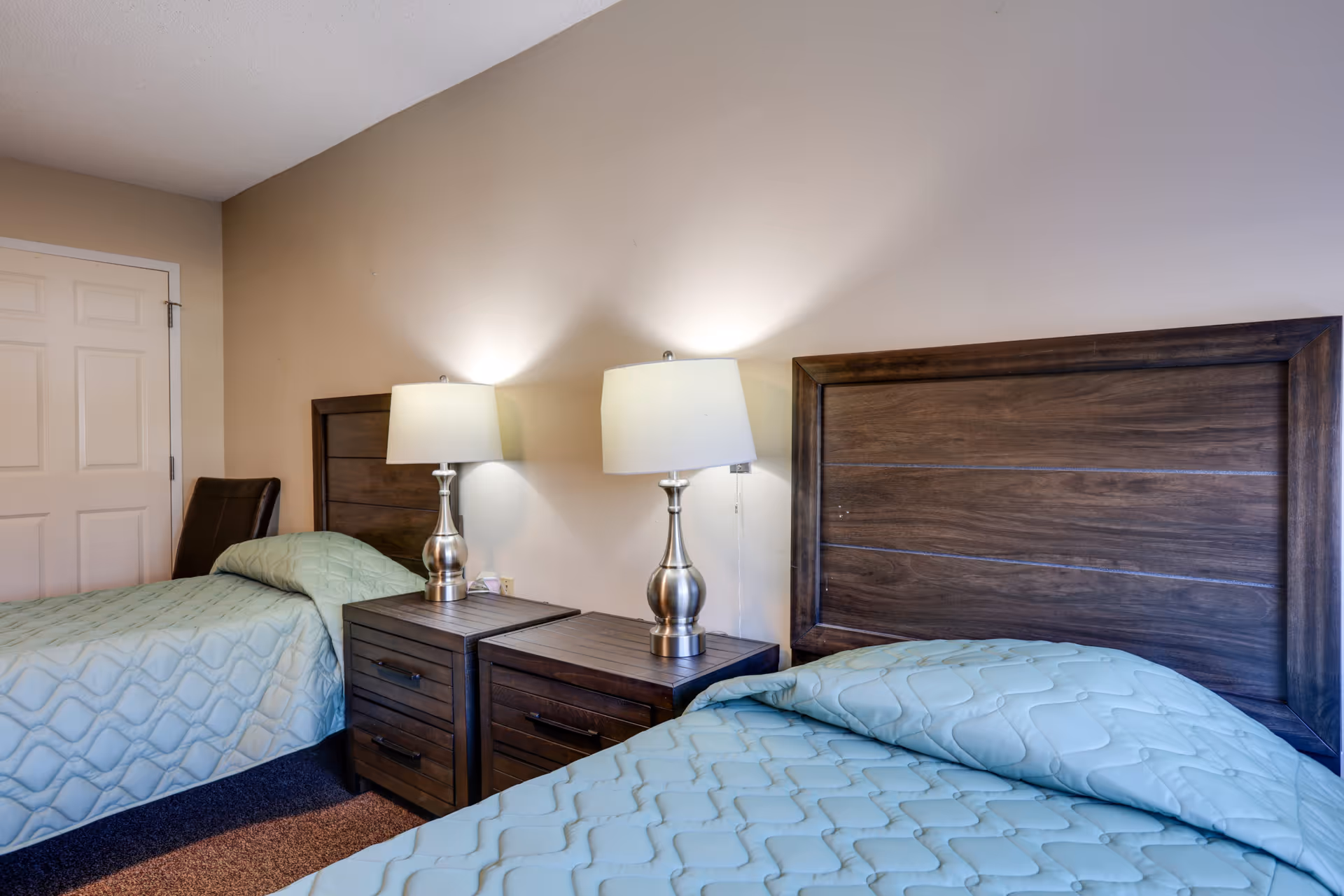 A senior living facility bedroom with two single beds featuring wooden headboards and light blue quilted bedspreads. Between the beds are two matching wooden nightstands, each with a silver lamp with a white lampshade. A closed white door and a dark brown chair are visible in the background.