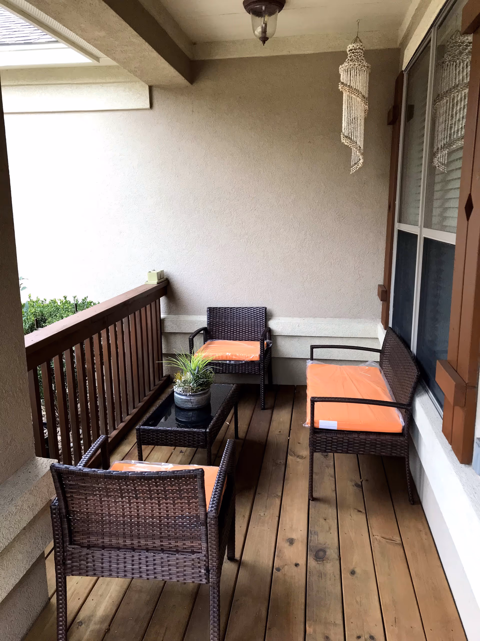A covered wooden porch with brown wicker furniture including two chairs, a loveseat, and a glass-top coffee table. The furniture has orange cushions still wrapped in plastic. A small potted plant is on the coffee table, and a hanging decorative wind chime is suspended from the ceiling near a window with brown shutters.