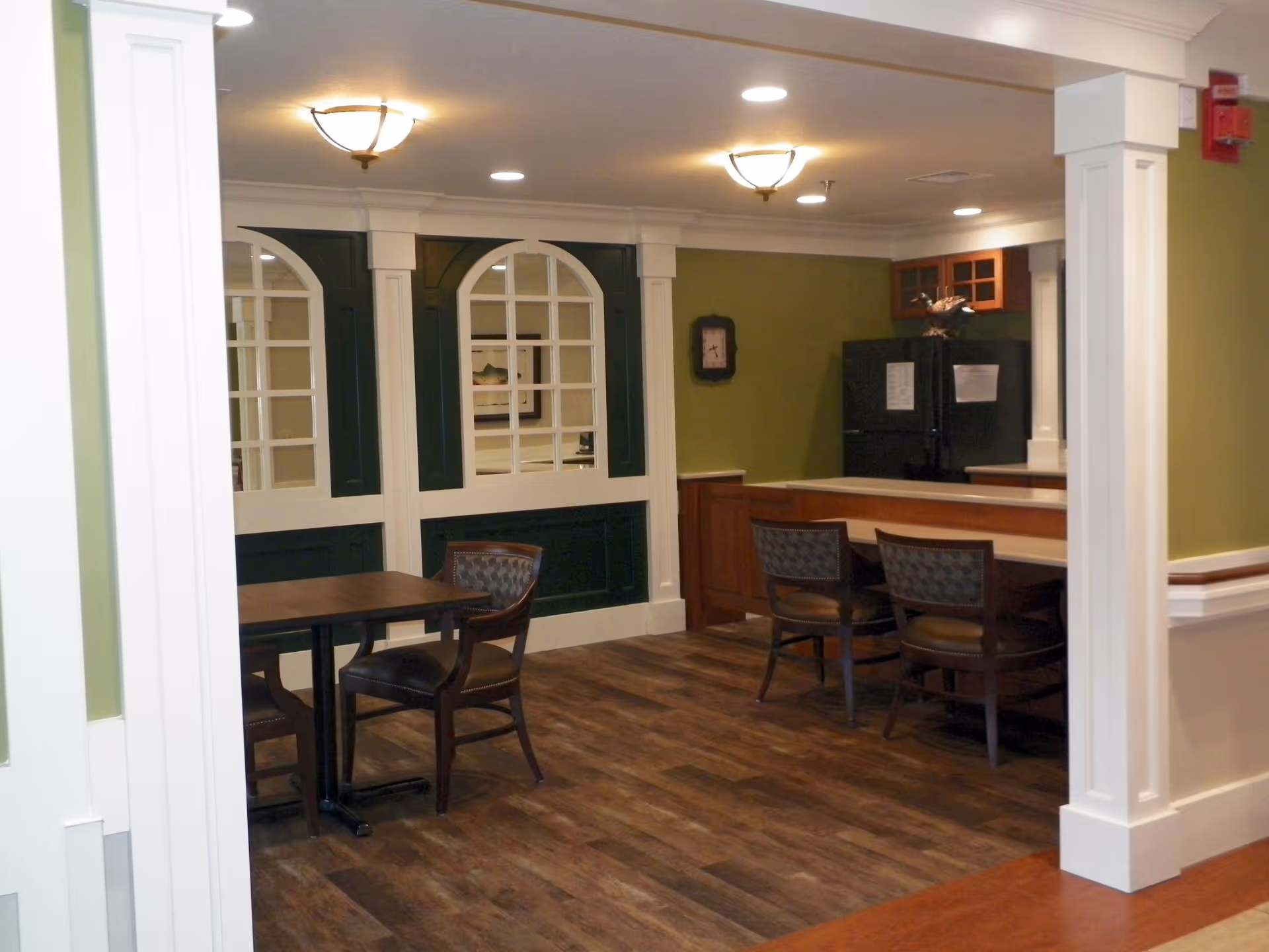 Interior view of a common area in a senior living facility featuring a small dining table with two chairs, a counter with three chairs, green and white walls with decorative window-like panels, wood flooring, and ceiling lights. A black refrigerator and a clock are visible in the background.