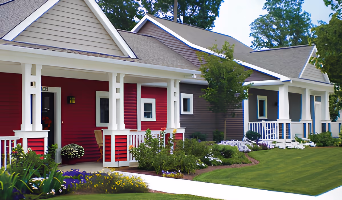 Exterior view of a residential building with a red and gray facade, white trim, and a covered porch. The building is surrounded by well-maintained green lawns, bushes, and flowering plants under a clear sky.