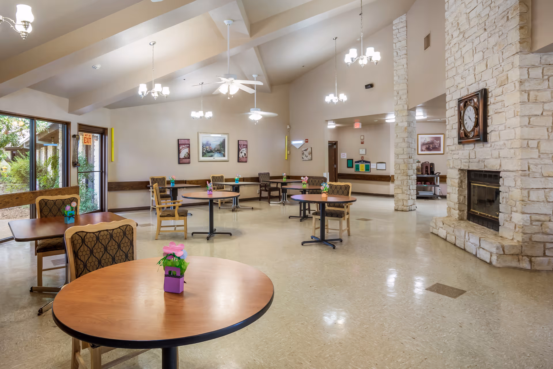 A spacious common area with round wooden tables and cushioned chairs arranged throughout the room. Each table has a small decorative flower pot. The room features a stone fireplace with a clock above it, beige walls, and several ceiling fans with lights. Large windows and a glass door provide natural light and a view of greenery outside.