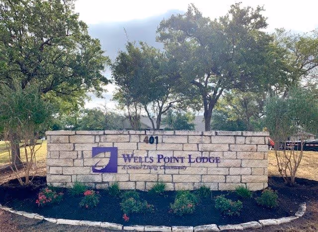 Stone sign for Wells Point Lodge, a senior living community, with the address number 401 on top. The sign is surrounded by landscaped plants and trees in the background under a partly cloudy sky.