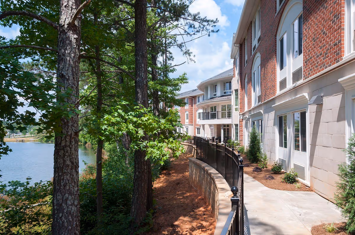 Outdoor view of The Georgian Lakeside facility showing a paved walkway along the side of a brick building with multiple windows. The walkway is bordered by a black metal fence and landscaping with small shrubs. On the left side, there are tall trees and a body of water visible under a partly cloudy sky.