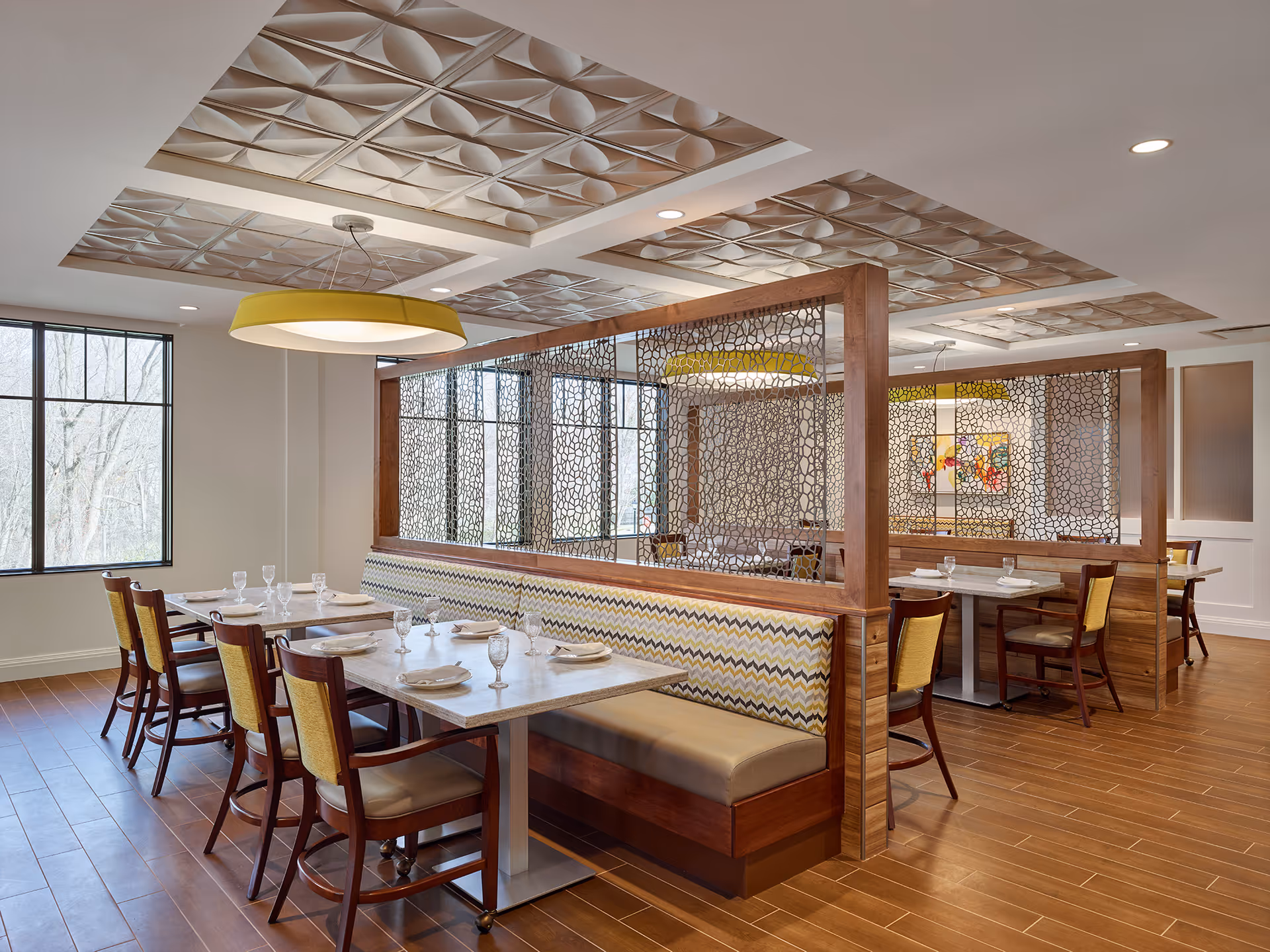 A modern dining area with wooden floors and a decorative ceiling. The space features a long cushioned bench with a zigzag patterned backrest, separated by wooden partitions with intricate metal designs. Several tables are set with plates, napkins, and glassware, surrounded by wooden chairs with yellow cushions. Large windows allow natural light to fill the room.