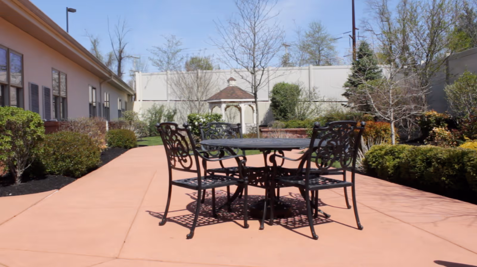 Outdoor patio area with a round metal table and four matching chairs on a paved surface, surrounded by bushes and trees with a small gazebo in the background.