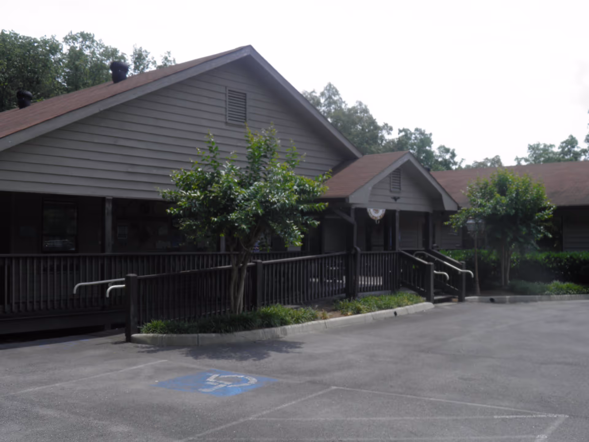 Exterior view of a single-story building with a brown roof and gray siding, featuring a wheelchair accessible ramp and stairs leading to the entrance. There are small trees and shrubs along the front, and a parking lot with a marked handicapped parking space in the foreground.