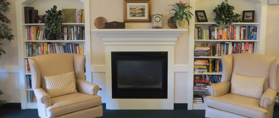 A cozy living room area featuring two beige wingback chairs with patterned cushions facing a white fireplace mantel. On either side of the fireplace are built-in bookshelves filled with books and decorative plants. The mantel is decorated with a framed picture, a clock, and small plants.