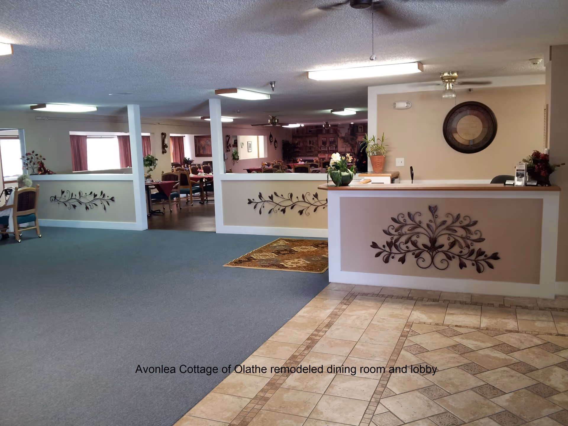 Interior view of a senior living facility lobby and dining room area. The foreground shows a reception desk with decorative metal wall art and a potted plant. The floor near the desk is tiled, transitioning to carpet in the lobby area. Beyond the half walls with decorative metal accents, there are tables and chairs arranged in the dining room with windows covered by curtains.