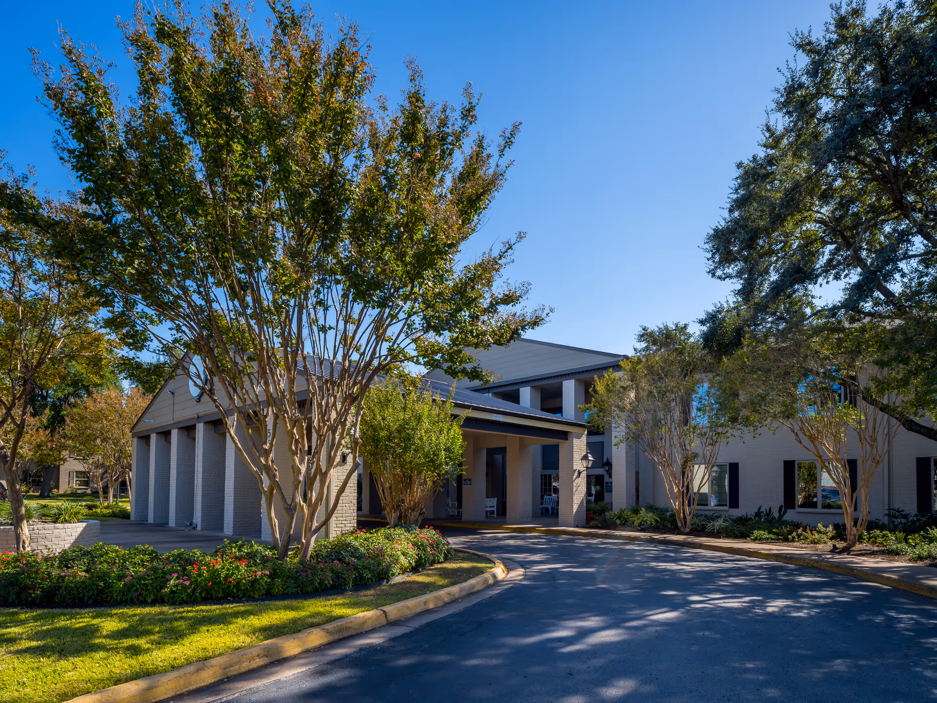 Exterior view of a senior living facility building with a covered entrance, surrounded by trees and landscaped greenery under a clear blue sky.