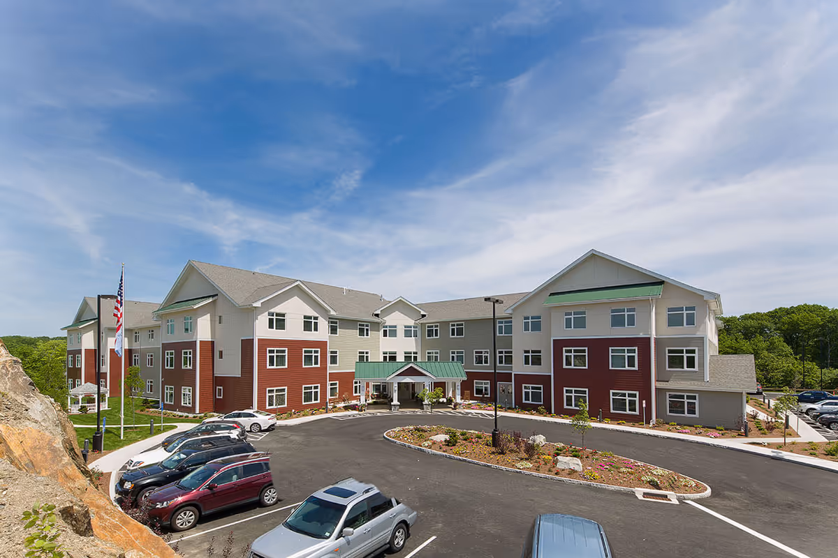 Exterior view of Benchmark Senior Living at Split Rock, a three-story building with a combination of beige and red siding, green awnings, and a covered entrance. Several cars are parked in the lot in front of the building, and there are landscaped areas with flowers and rocks. The sky is clear with some clouds.