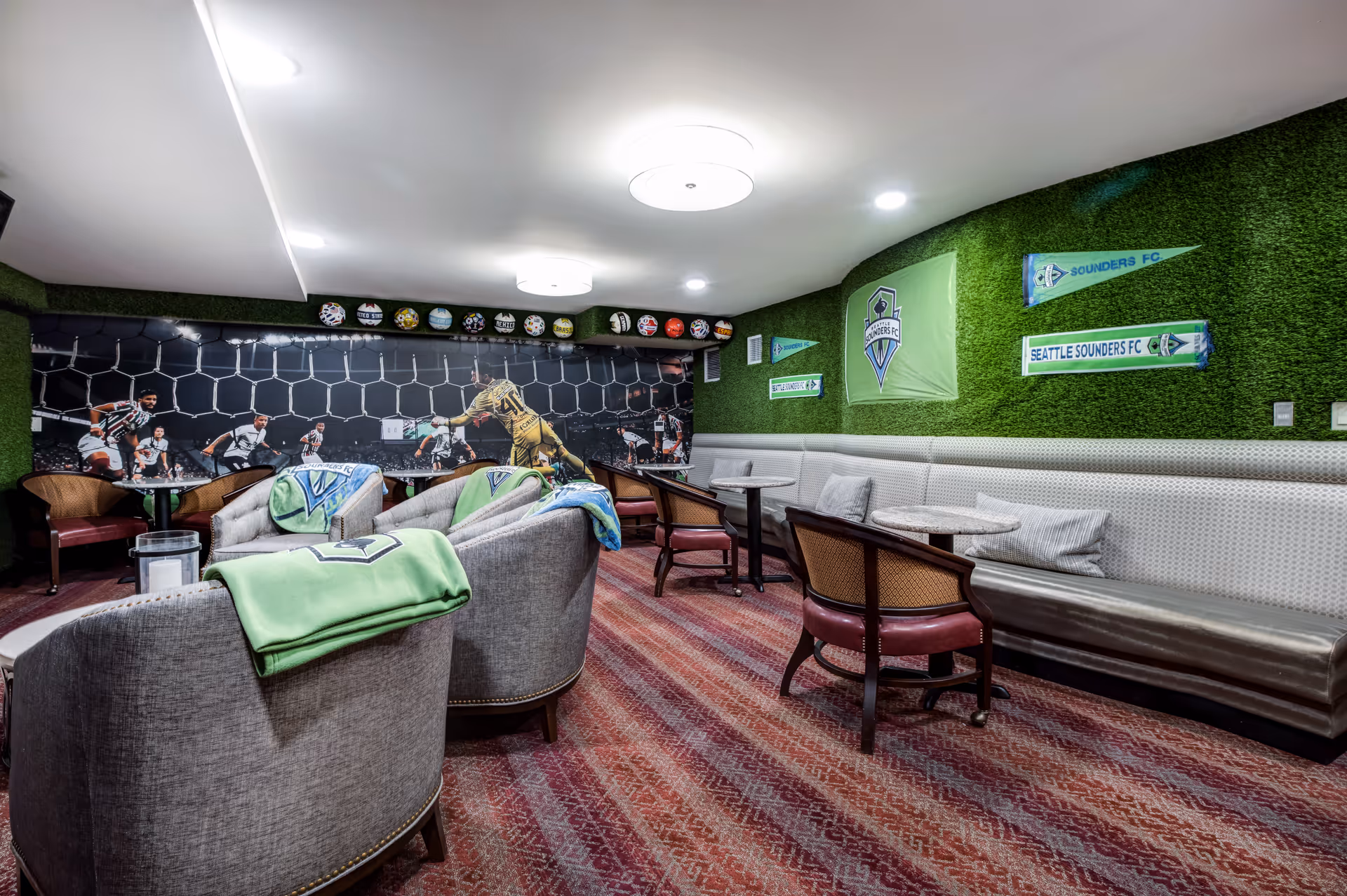 A cozy lounge area decorated with Seattle Sounders FC soccer memorabilia including flags, scarves, and a large mural of a soccer game with a goalkeeper diving to make a save. The room features comfortable armchairs with green blankets draped over them, small round tables, and a long cushioned bench along one wall with pillows. The walls have green turf-like covering and the floor is carpeted in a red patterned design.