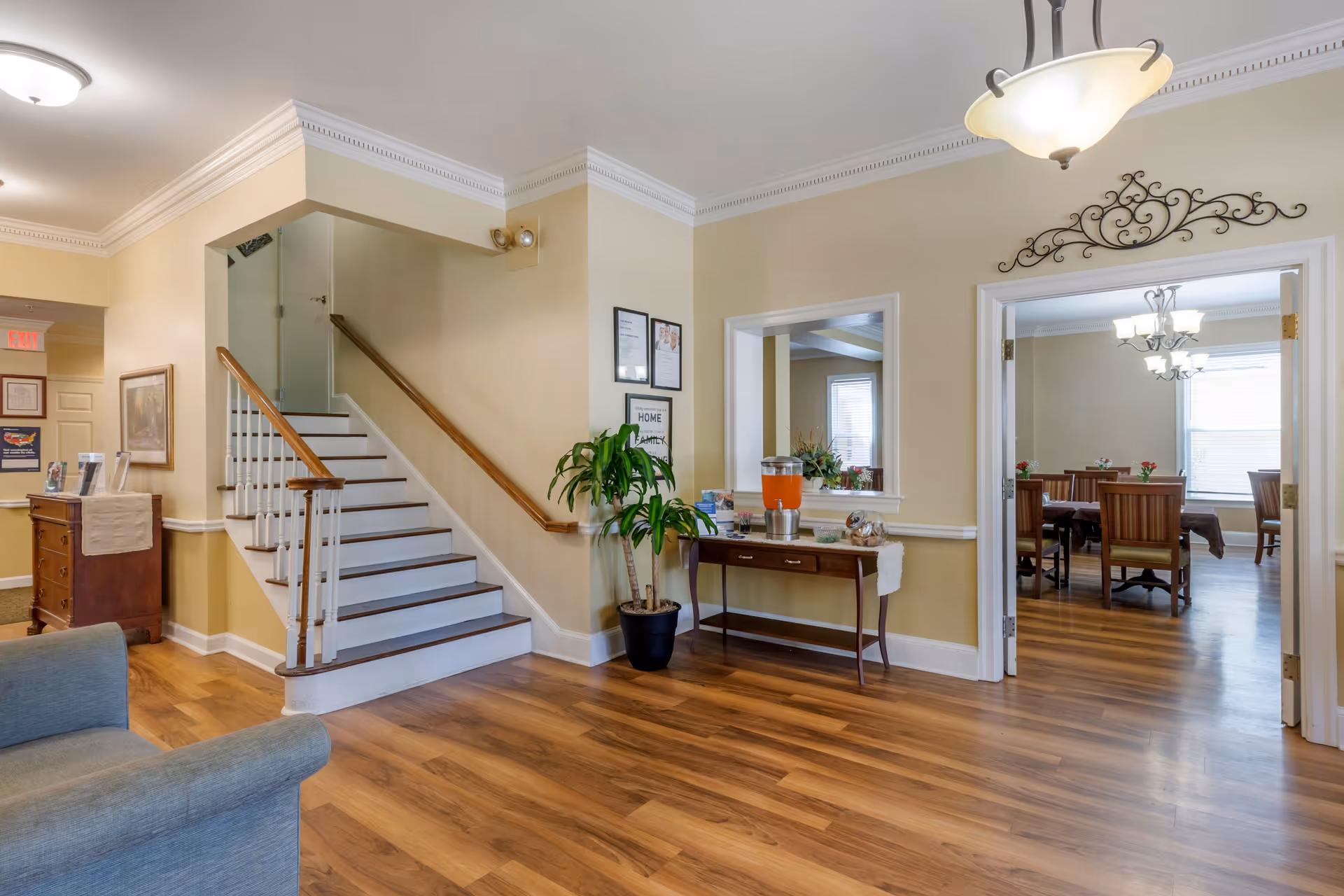 Interior view of a senior living facility showing a hallway with wooden flooring, a staircase with white steps and wooden handrails, a small table with a beverage dispenser and snacks, a potted plant, and an adjacent dining room with tables and chairs visible through an open doorway.