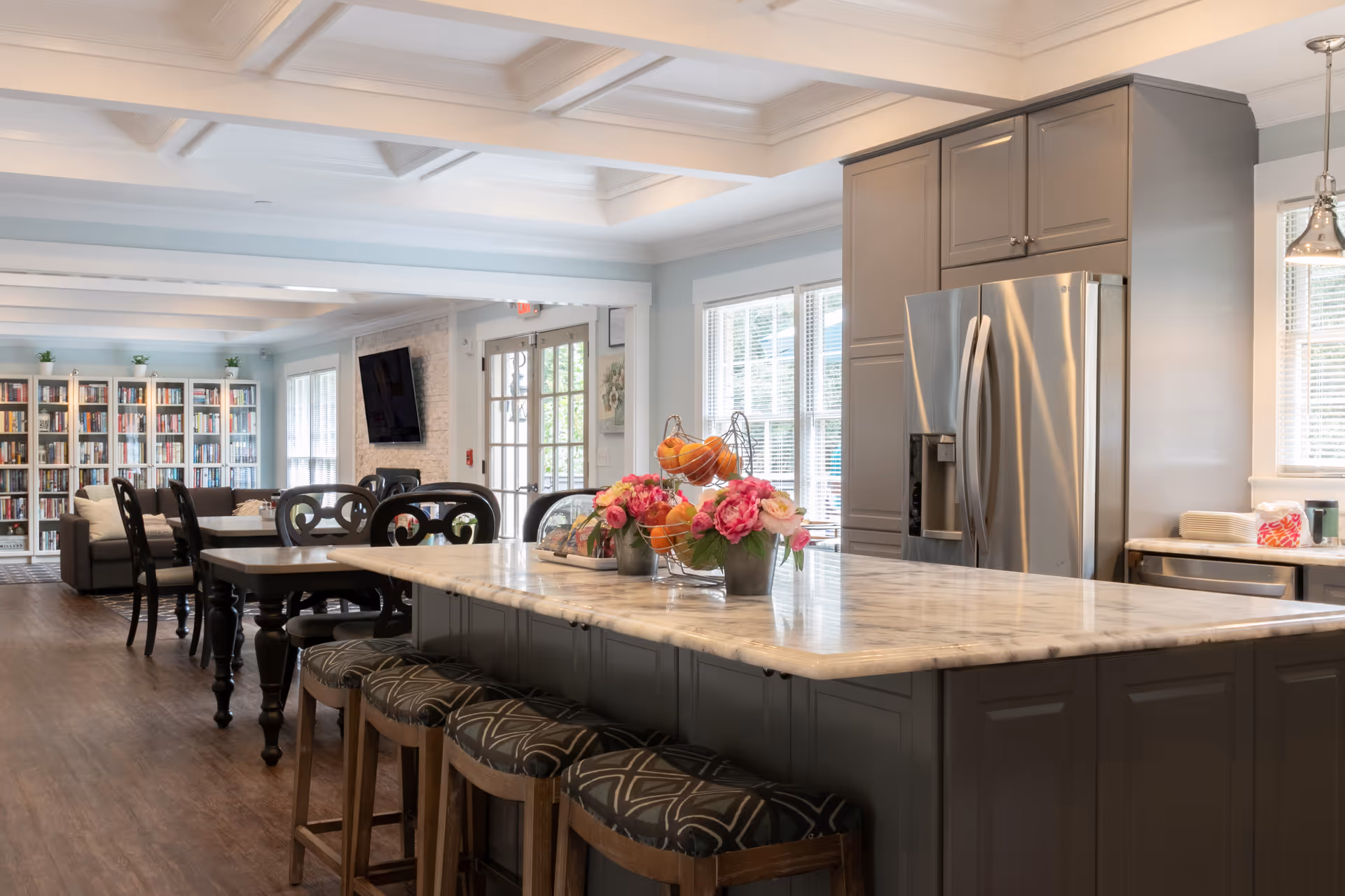 A spacious and bright senior living facility kitchen and common area with a large marble island countertop surrounded by four cushioned stools. The kitchen features gray cabinets and a stainless steel refrigerator. In the background, there is a dining area with tables and chairs, a bookshelf filled with books, a wall-mounted TV, and large windows letting in natural light.