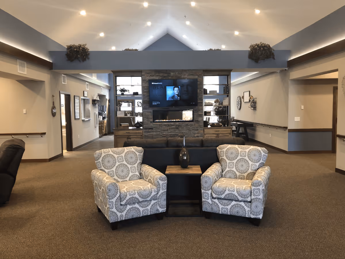 A spacious and well-lit living room area with two patterned armchairs facing a black sofa and a wooden side table with a decorative vase. In the background, there is a stone fireplace with a mounted flat-screen TV above it, flanked by built-in shelves with decorative items. The room has high vaulted ceilings with recessed lighting and neutral-colored walls and carpet.