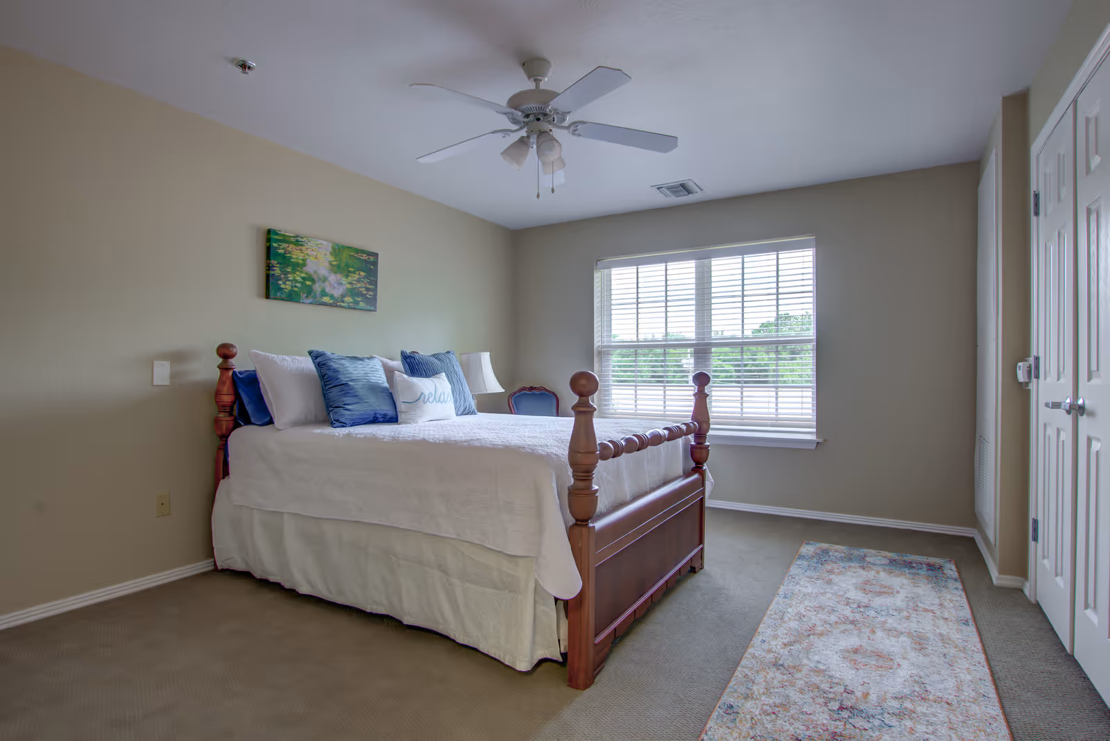 A bright bedroom with a wooden bed frame, white bedding, and several blue and white pillows. There is a ceiling fan above, a window with blinds letting in natural light, a small chair in the corner, a lamp on a nightstand, a colorful rug on the carpeted floor, and a painting on the beige wall.