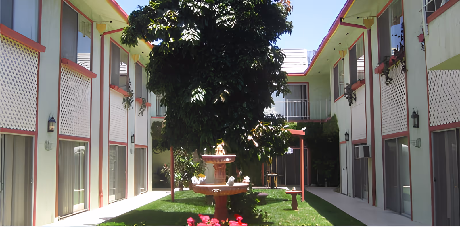 Outdoor courtyard area of a retirement facility with a central tree and a tiered water fountain surrounded by green grass. The courtyard is flanked by two-story buildings with balconies and sliding glass doors, featuring white lattice panels and red trim.