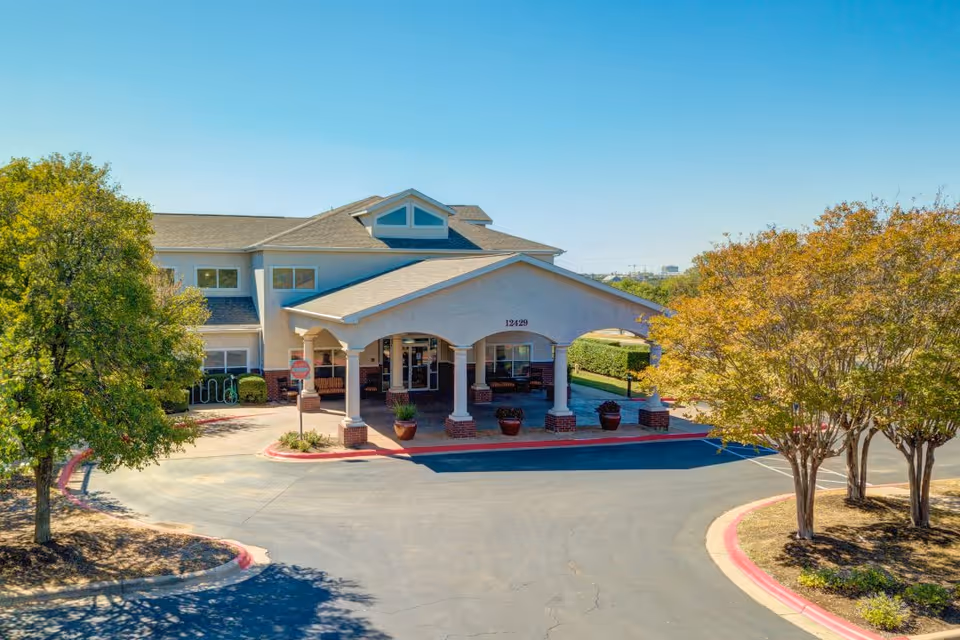 Front exterior view of a two-story senior living facility building with a covered entrance supported by columns. The building is surrounded by trees and landscaping under a clear blue sky.