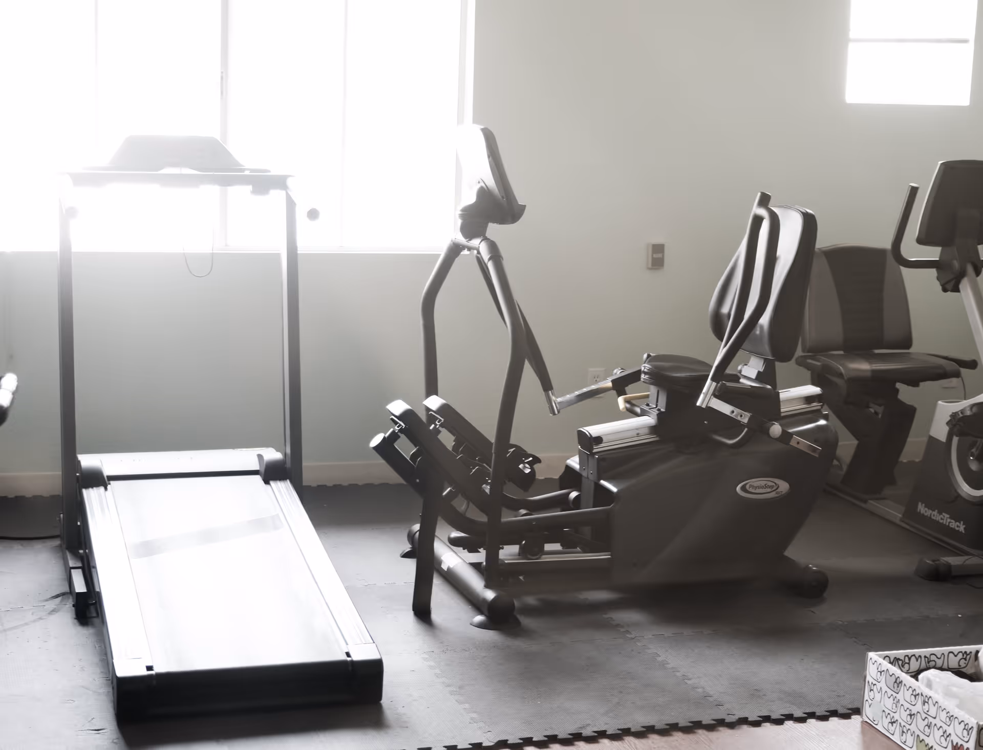 A small exercise room with a treadmill, a recumbent elliptical machine, and a recumbent bike. The room has light-colored walls and a window letting in natural light.