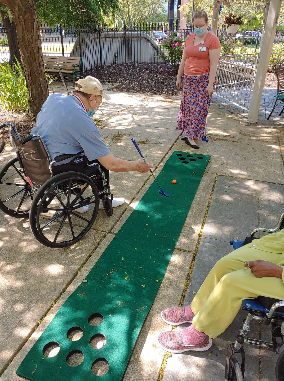 An elderly man in a wheelchair playing mini golf on a green putting mat outdoors, while a woman wearing a mask and a colorful skirt watches. Another person in a wheelchair wearing yellow pants and pink shoes is partially visible on the right side. The setting is a shaded patio area with trees and plants nearby.