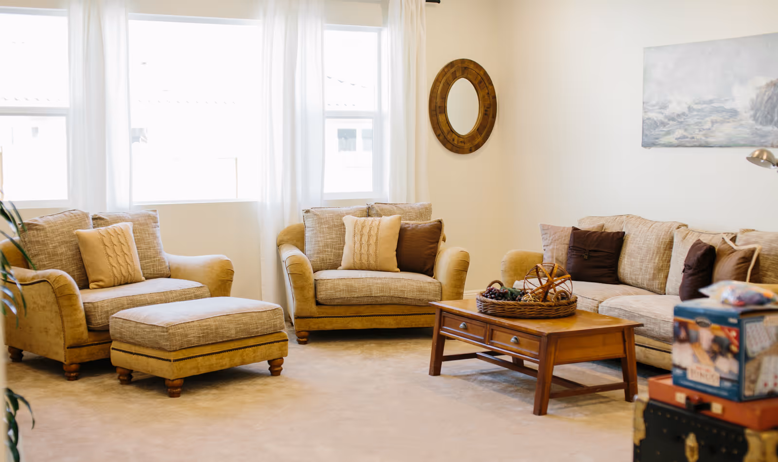 A cozy living room with beige upholstered armchairs, a matching ottoman, and a sofa adorned with various throw pillows. A wooden coffee table with decorative items sits in the center. The room has light-colored walls, two large windows with white curtains, a round wooden-framed mirror on the wall, and a painting depicting a seascape.