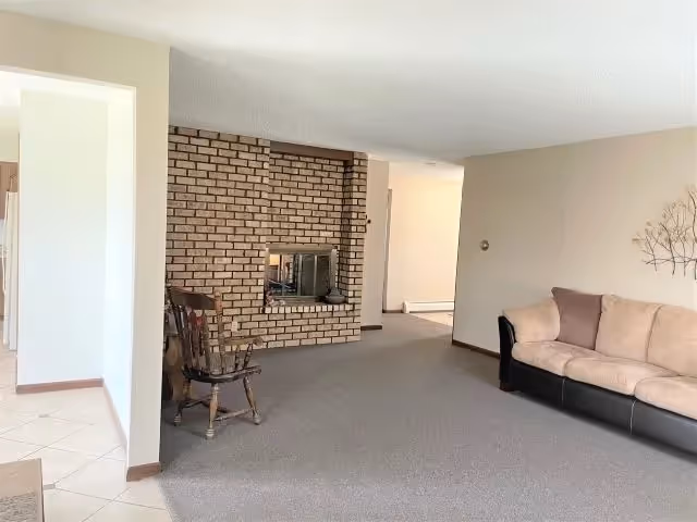 A living room area with a beige and black sofa on the right, a wooden rocking chair near a brick fireplace in the center, and beige walls with a decorative metal wall art. The floor is carpeted in the living area and tiled near the entrance to another room.