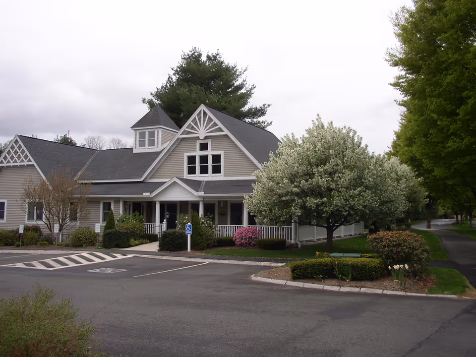A two-story, house-style retirement community building with a front porch, landscaped shrubs, and a blooming tree beside a parking area.