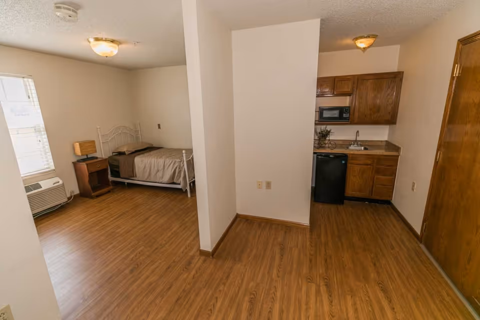 Interior view of a senior living facility room with a single bed, nightstand with a lamp, window with blinds, and an air conditioning unit on the left side. On the right side, there is a small kitchenette with wooden cabinets, a microwave, a sink, and a mini refrigerator. The floor is wood laminate, and the walls are painted white.