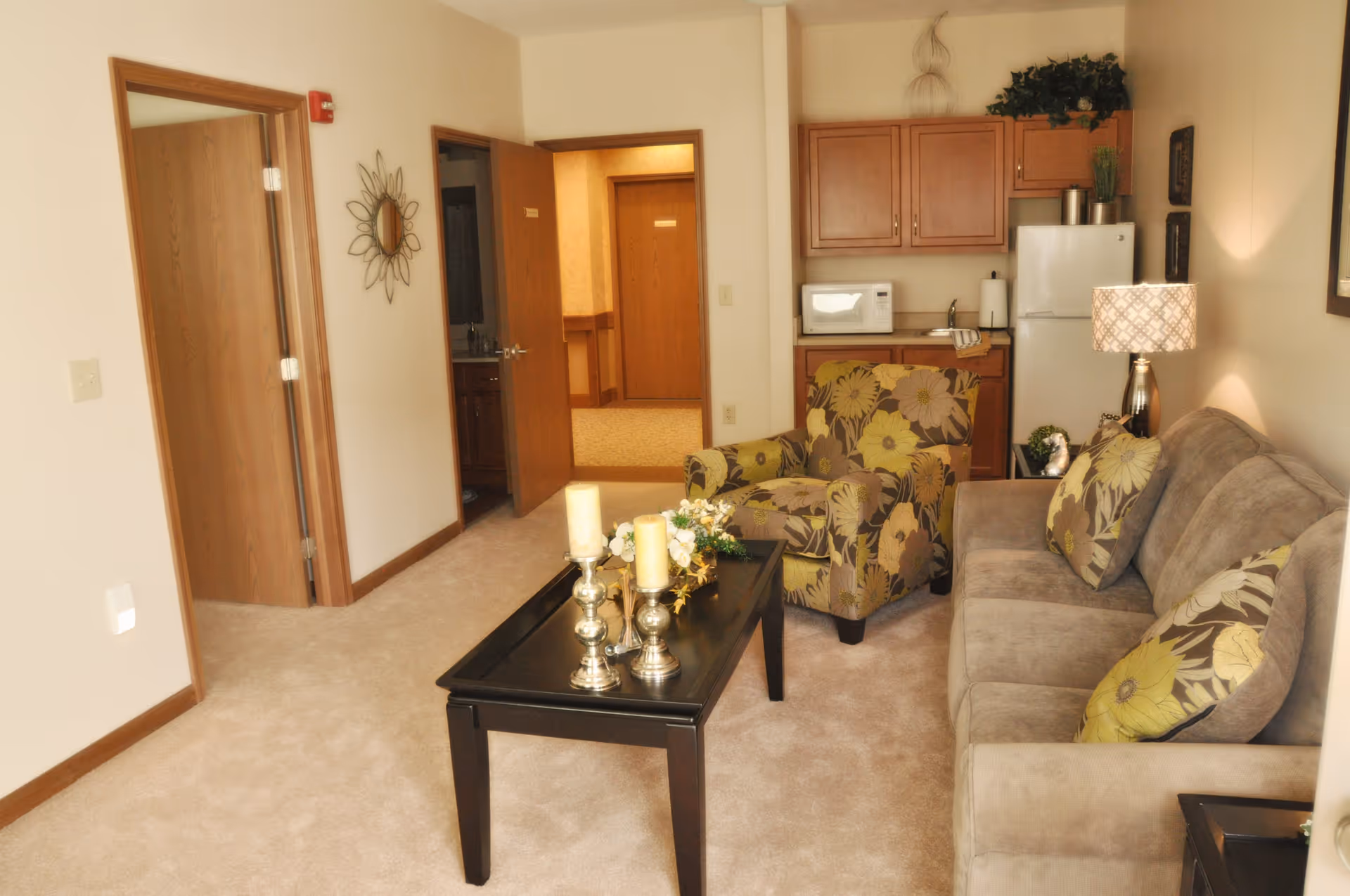 A cozy living room area in a senior living facility featuring a beige sofa with floral cushions, a matching floral armchair, a black coffee table with decorative candles and flowers, a lamp on a side table, and a small kitchenette with wooden cabinets, a microwave, and a refrigerator in the background.