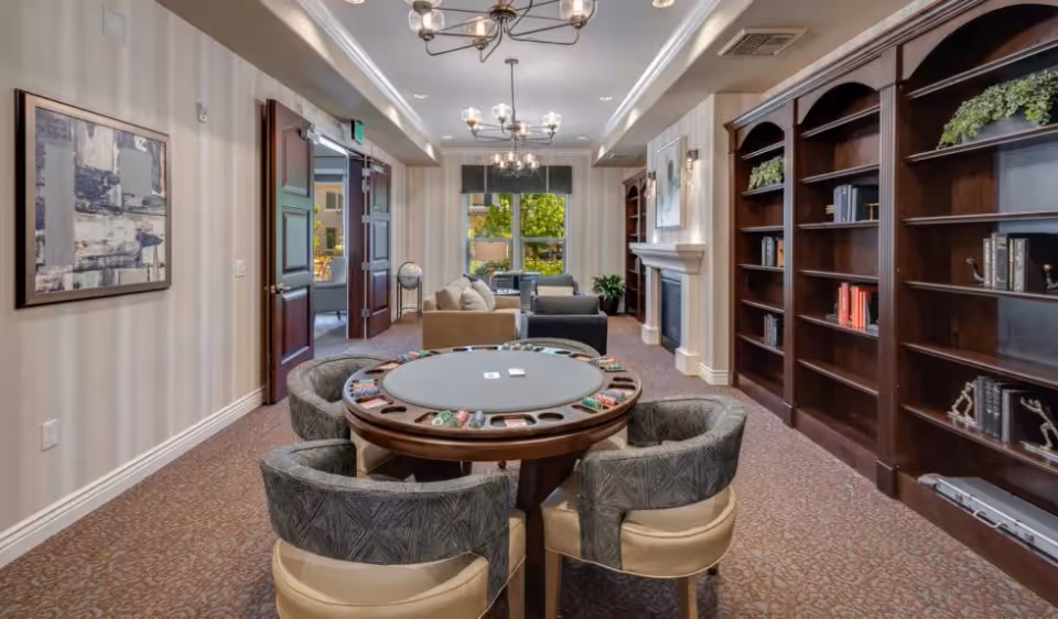 A cozy senior living common area featuring a round poker table with four cushioned chairs in the foreground. The room has beige walls with subtle stripes, a carpeted floor, and built-in dark wood bookshelves filled with books and decorative plants on the right. In the background, there are two sofas facing each other near a fireplace with a painting above it. Large windows let in natural light, and elegant chandeliers hang from the ceiling.