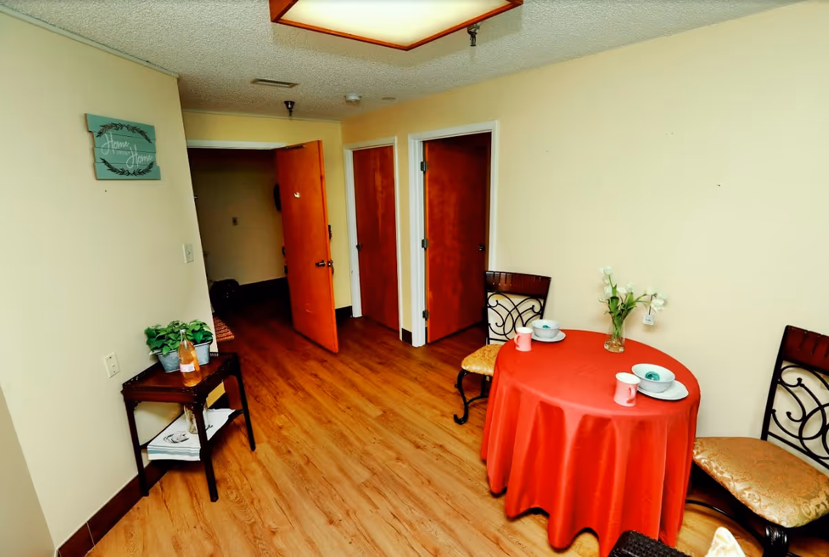 Interior view of a senior living facility room with wooden flooring, beige walls, and three wooden doors. A small round table covered with a red tablecloth is set with two place settings and a vase of white flowers. Two chairs with patterned cushions are placed around the table. A small side table with a plant and a bottle is against the left wall, which has a decorative sign that reads 'Home Sweet Home'.