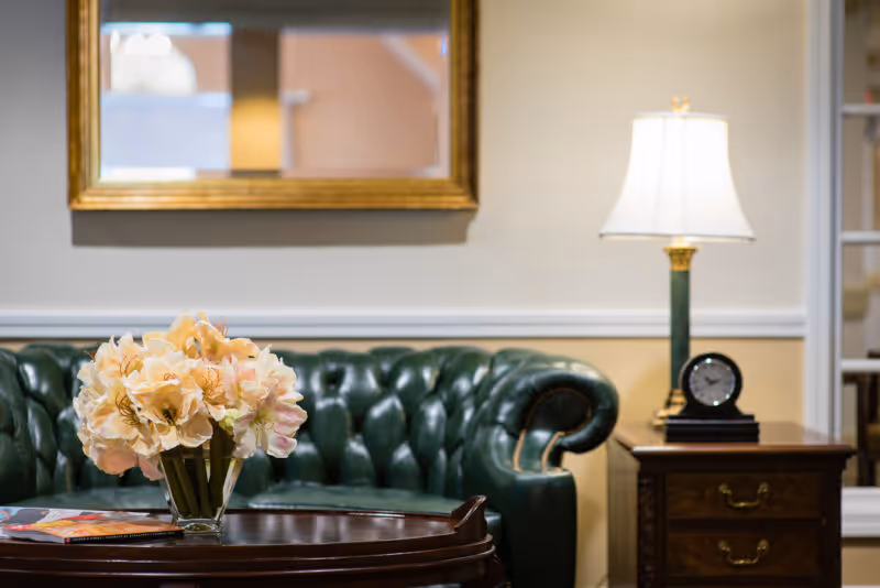 A cozy living room area featuring a dark green tufted leather sofa, a wooden coffee table with a glass vase of light pink flowers, a wooden side table with a classic black clock and a white lampshade on a lamp, and a gold-framed mirror hanging on the beige wall.