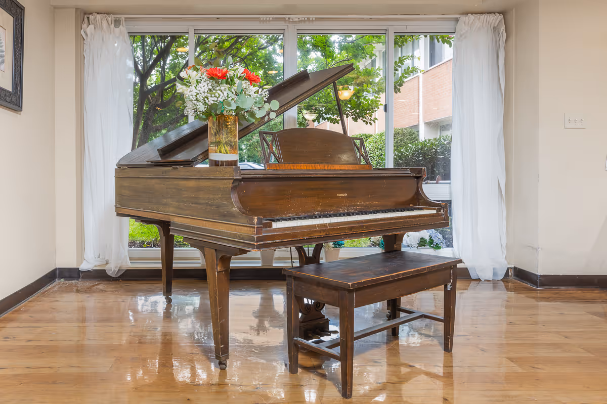 A wooden grand piano with an open lid and a matching wooden bench is placed on a polished wooden floor in front of large windows with white curtains. A vase with red and white flowers sits on top of the piano. Outside the window, green trees and a brick building are visible.